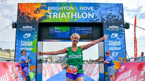 A participant is celebrating after passing the finish line at the Brighton and Hove Triathlon. They are wearing a green Macmillan branded vest, a event number card, and shorts. People are cheering on either side of the participant. 