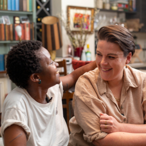 Zizi with their partner. They are sitting inside a house at a table. Zizi is wearing a white top. Their partner has on a light brown top.