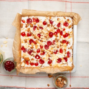 A view of a cherry and almond tray bake from above. The tray bake is on a table that has a white and red chequered table cloth on it. Next to the tray bake are jars with items like almonds and cherries. 