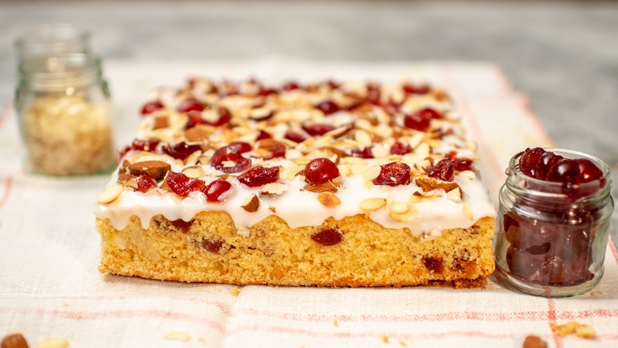 A golden-brown cherry and almond tray bake topped with smooth white icing, glistening glazed cherries, and sliced almonds. Presented on a striped cloth with jars of extra cherries and almonds beside it,
