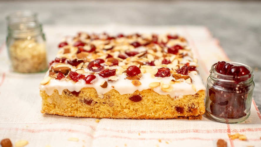 A cherry and almond tray bake is on a white and red patterned table cloth. The tray bake is topped with cherries and almonds. Next to it are jars with almonds and cherries, and on the table cloth are almonds. 