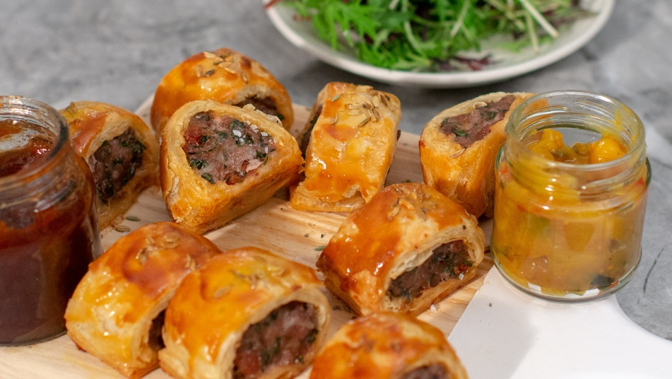 Several chorizo sausage rolls are on a wood and plastic board. They are jars of condiments also on the board. Behind them is a plate with different leaf vegetables. 