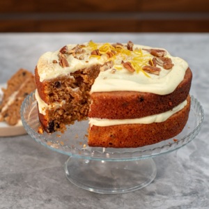 A moist, two-layer carrot cake with creamy white frosting between the layers and on top, garnished with chopped pecans and orange zest. A slice has been cut to reveal the rich interior speckled with carrot and raisins, all presented on a glass stand.