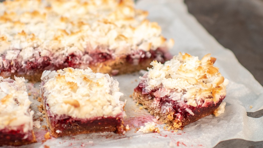 A freshly baked dessert bar with a light crust, vibrant red berry filling, and toasted coconut flakes sits on parchment paper. One piece is cut out, revealing its moist, crumbly layers—an irresistibly sweet and fruity treat.