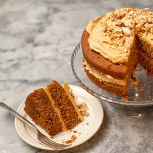A two layered coffee, walnut and cardamom cake is on a glass cake stand. Next to the stand is a slice of the cake on a white plate with a silver fork. The cake is topped with a caramel coloured frosting and crushed walnuts. 