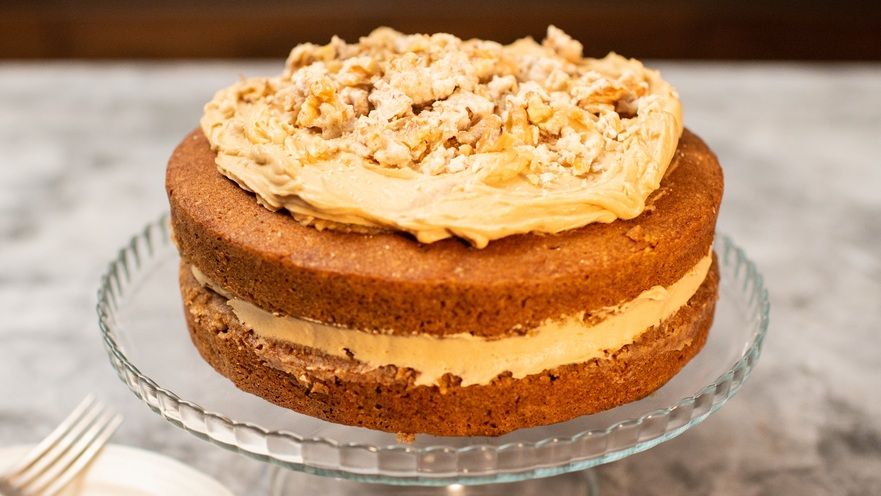 A two-layered sponge cake with a golden-brown crumb and creamy frosting between the layers and on top, garnished with chopped walnuts. Presented on a glass cake stand.