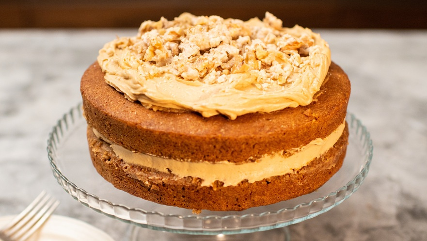 A two layered coffee, walnut and cardamom cake is on a glass cat stand. The cake is topped with caramel coloured frosting and crushed walnuts. Next to the stand is a white plate with a silver fork on it. 