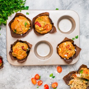 A view of muffins from above. Some muffins are placed in a muffin tin, and some are on the surface where the tin is placed. The muffins are wrapped in brown parchment paper. Next to the muffins and tray is a bundle of parsley and some cut up tomatoes. 