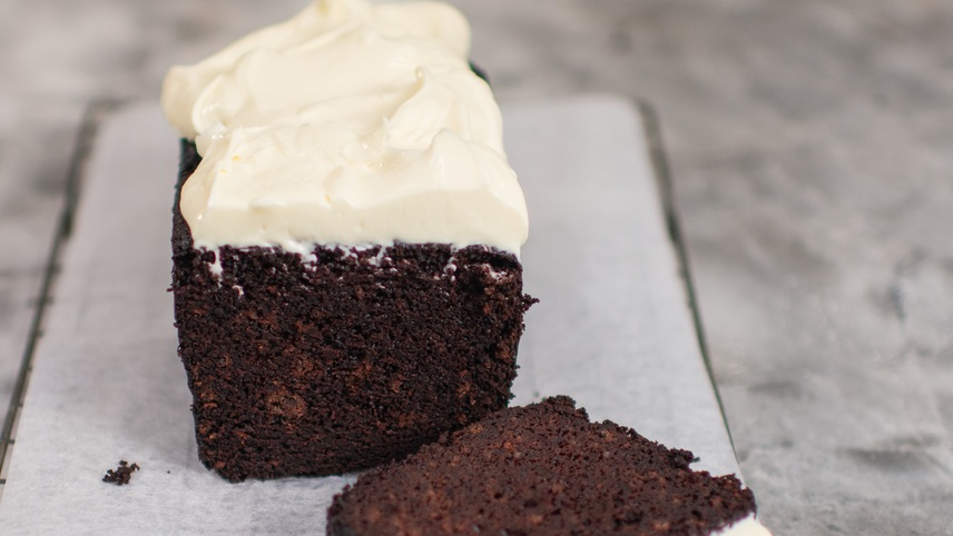 A rich, moist chocolate loaf cake topped with a thick layer of creamy white frosting, resting on parchment paper. A single slice lies in front, revealing its dense, decadent texture.