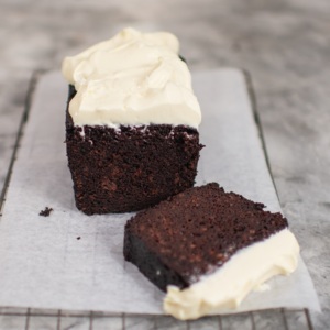 A rich, moist chocolate loaf cake topped with a thick layer of creamy white frosting, resting on parchment paper. A single slice lies in front, revealing its dense, decadent texture.