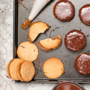 A baking tray filled with round, golden cookies—some topped with glossy chocolate. A piping bag oozes rich caramel in the corner, white crumbled cookie pieces reveal a soft, crumbly texture.