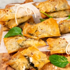 Several spinach and feta parcels are on a wooden board. Some of the parcels have baking parchment and string wrapped around them. Also scattered on the board are spinach leaves. 