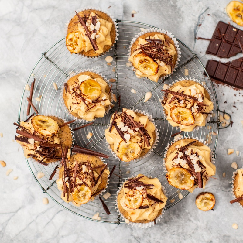 Several vegan peanut butter and banana cupcakes are on a cooling rack. The top of the cupcakes are decorated with frosting, chocolate flakes, and baked banana pieces. Next to the cupcakes is a broken bar of chocolate. 