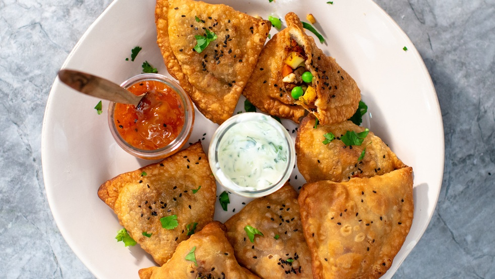 Several vegetable samosas are on a white plate. There is also a jar of a white sauce and a chutney on the plate. The chutney jar has a utensil sticking out of it. 