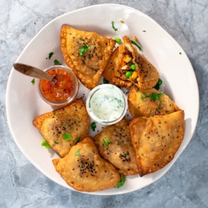 Several vegetable samosas are on a white plate. There is also a jar of a white sauce and a chutney on the plate. The chutney jar has a utensil sticking out of it. 