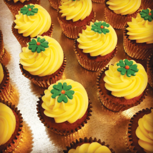 Rows of carrot cupcakes in metallic gold wrappers, each topped with a generous swirl of creamy yellow frosting and a delicate green fondant flower with a red center.