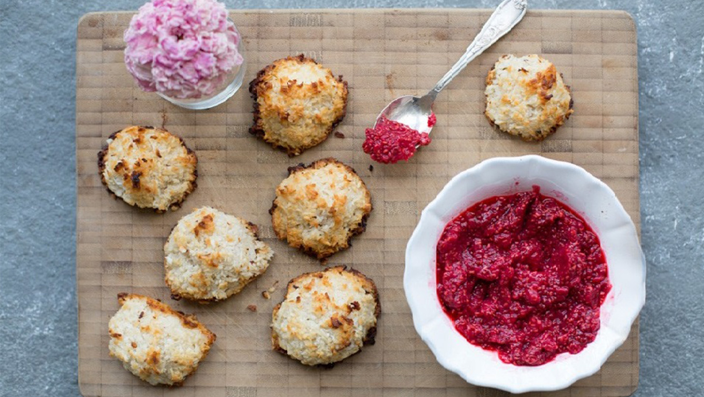 Several coconut macaroons are on a wooden cutting board. There is a white bowl with red chia seed jam in the bowl. There is also a silver spoon on the board that has a scope of the jam on it. There is also a glass vase with a pink flower in it. 