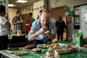 A man is leaning over a Coffee Morning table and taking a bite from a pastry. The man has on a blue shirt. The table has many different desserts and other treats on it. Behind the man are other people eating food and socialising. 