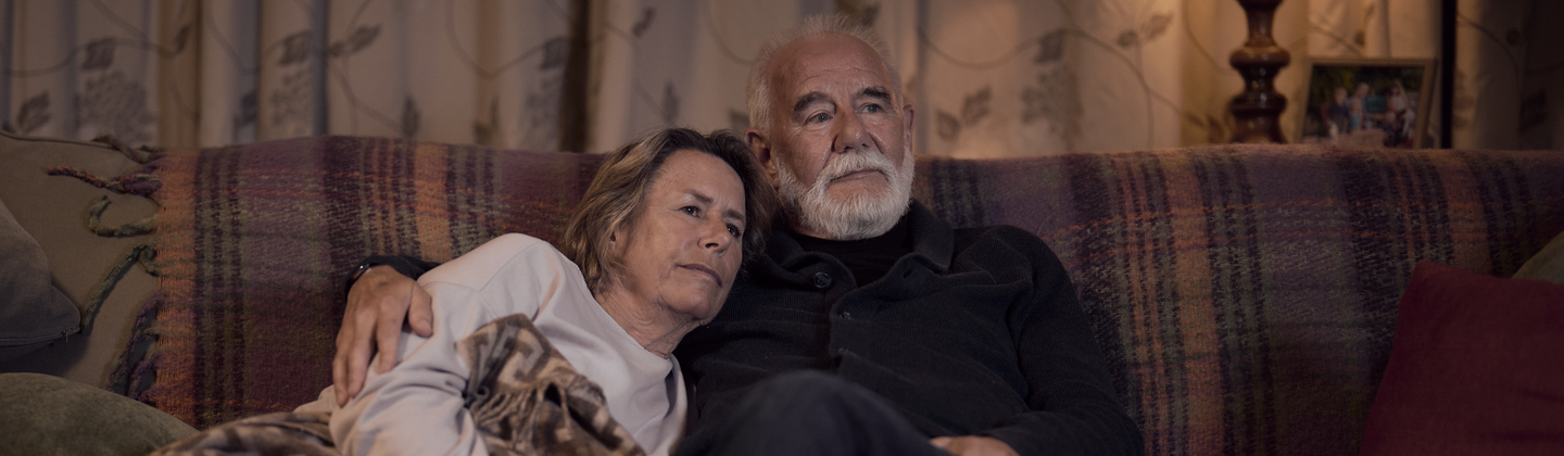 A white women and man sit on a sofa under a blanket comforting each other.