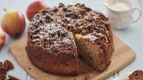 An apple cake with pecan nut streusel is resting on a wooden board. The cake has been sliced and a piece is missing. Around the cake is scattered pecans, apples, and a small white jug with a white cream in it. 