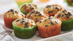 Many muffins are on a white plate. The muffins are in green or orange muffin liners. The muffins are topped with seeds. 