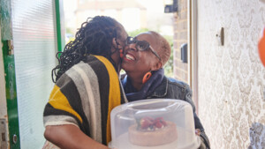 Two people are standing in a doorway for a home. The are hugging one another. One person is holding a cake container that has a cake decorated with fruit on it. 
