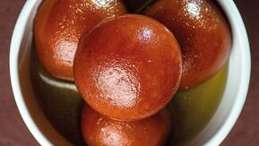 Four golden-brown spherical gulab jamuns glistening in sugar syrup. Their glossy, syrup-soaked surface and rich color make them look irresistibly moist, sweet, and indulgent.