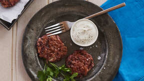 Two beetroot latkes are on a metallic plate. Next to them is a garnish, a pot with a white cream, and a silver fork. The plate is on top of a dark blue cotton napkin and a wooden table. There is also a baking tray with two latkes next to the plate. 