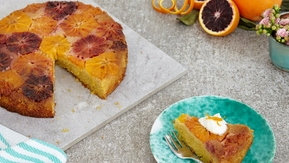 A polenta cake is on top of a square marble plate. A slice has been cut out of the cake. The slice is on a blue patterned plate. It has been topped with cream and an orange garnish. The plate also has a silver fork on it. 