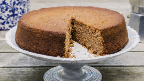 A chestnut cake is on top of a white cake stand. A slice has been cut from the cake. The cake stand is on top of a wooden table. Behind it is a white and blue plant pot and a silver stove top espresso coffee maker. In front of the cake stand is a plate with the cake slice on it. 
