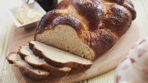 A loaf of challah bread is resting on top of a wooden serving board. Part of the loaf has been cut in slices. Also on the wooden board is an open jar with a spoon resting on top of it and a small plate with butter in it. 