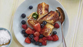 Two cheese blintzes are on a light blue plate. One of the blintzes has been sliced in half. They are topped with strawberries and blueberries, and a white dusting. There is also a silver spoon on the plate. 