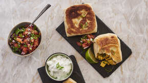 Two dark rectangular slate plates are on top of a light marble table top. On the plates are rotis. On one plate a roti has had a bite taken out of it. Next to the plates is a small bowl with chopped vegetables that has a spoon in it. One plate also has a pot of a white dip in it. 