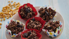 Six rich chocolate and dried fruit clusters nestled in vibrant cupcake liners—red, floral, and polka-dotted—sit on a plate. Packed with oats, chocolate, and chewy dried fruits, their textured surface looks indulgent and satisfying.