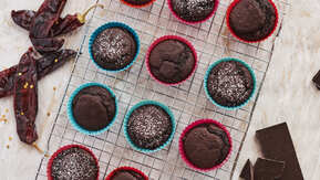 Many muffins are on top of a metal baking cooling rack. The cupcakes are in blue or red cupcake liners. Some of the cupcakes have a sprinkle of white powder on top. Next to the cooling rack are a few red chillies and a broken bar of chocolate. 