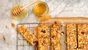 A batch of freshly baked granola bars packed with oats, nuts, seeds, and dried fruits rests on a cooling rack. Rectangular pieces are cut and ready to serve, while a jar of golden honey with a dripping dipper adds a glossy, indulgent touch.