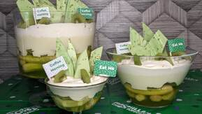 Three dessert bowls layered with fresh kiwi slices and creamy white mousse, topped with green chocolate shards and playful flags reading “Coffee Morning” and “Eat Me.” The vibrant layers and whimsical garnishes make these treats look irresistibly fresh and inviting.