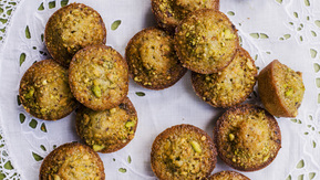 A batch of freshly baked pistachio mini muffins with golden-brown crusts and crunchy green pistachio pieces, arranged on a white lace doily. Set against a green tablecloth, the close-up view makes these nutty treats look irresistibly warm and inviting.