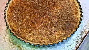 A freshly baked tart with a golden-brown crust and a smooth, speckled filling, lightly dusted with nutmeg. Nestled in a fluted-edge pan, the tart’s warm, aromatic appearance makes it look irresistibly comforting and delicious.