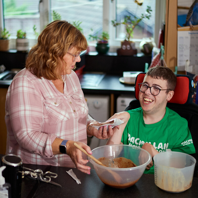 Two people are mixing ingredients in a clear bowl together in a kitchen. The person on the left has a white and pink chequered top and short brown hair. The person on the right is sitting on a mobility chair. They have short brown hair and are wearing a green Macmillan t-shirt.