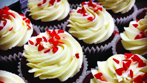 A close up view of many red velvet cupcakes lined up in rows. The cupcakes have white frosting and red sprinkles. The cupcakes are in white cupcake liners. They have a red sponge. 