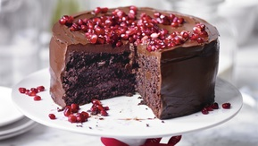 A chocolate and pomegranate layered cake is on top of a white cake stand. The cake has three layers and has chocolate frosting. It is topped with pomegranate seeds. The base of the cake stand has a red ribbon tied around it. 