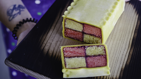 A Battenberg cake loaf is on a wooden board. A person wearing a blue apron with polka dots is holding the wooden board. The slice of cake has been cut from the cake. 