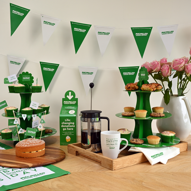 A table is decorated for a Coffee Morning event. There are cup cake stands, a French press, a tea pot, a vase with pink flowers and a small cake on a wood board. There is Macmillan branded decorations on the table, and bunting on the wall behind the table.