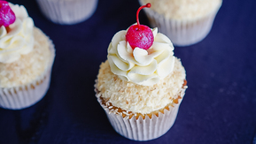 Four cupcakes are on placed on a black board that is being held by a person wearing a blue with colourful polka dots apron. The cupcakes are topped with white frosting and a red cherry. The cupcakes are in white cupcake liners. 