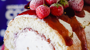 A close up view of an artic roll. The roll is topped with a strawberry jam and strawberries. Inside the roll is white ice cream. 