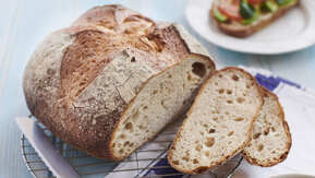 A sourdough loaf is on top of a metal cooling rack. The loaf has been cut into slices. Rest on the cooling rack is a metal knife. Underneath the cooling rack is a cotton napkin. Near the loaf is a plate with a slice of bread on it that has been topped with vegetables, and a bowl with a sliced avocado and sliced tomatoes. 