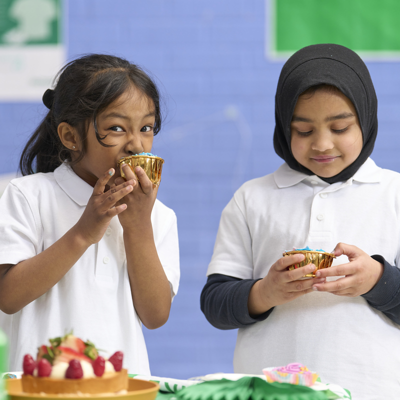 Two young people in school uniforms are holding cupcakes. The person on the left is biting into the cupcake. They appear to be in a school. 