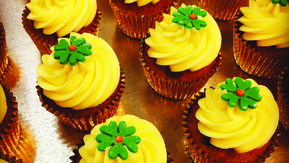Many cupcakes are lined up in rows. The cupcakes have yellow frosting and are topped with a green flower sprinkle. The cupcakes are in gold metal cupcake lining. 