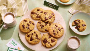A plate of golden-brown chocolate chip cookies with gooey melted chunks sits on a light green wooden table. Two frothy coffees in white and beige mugs, partially eaten cookies on side plates, and green-striped napkins create a cozy, inviting scene.